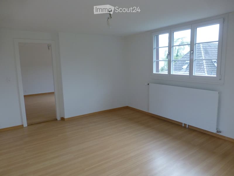 Empty room with wooden flooring, white walls, and large windows letting in natural light. A radiator is visible under the windows, and a door is seen on the left side.