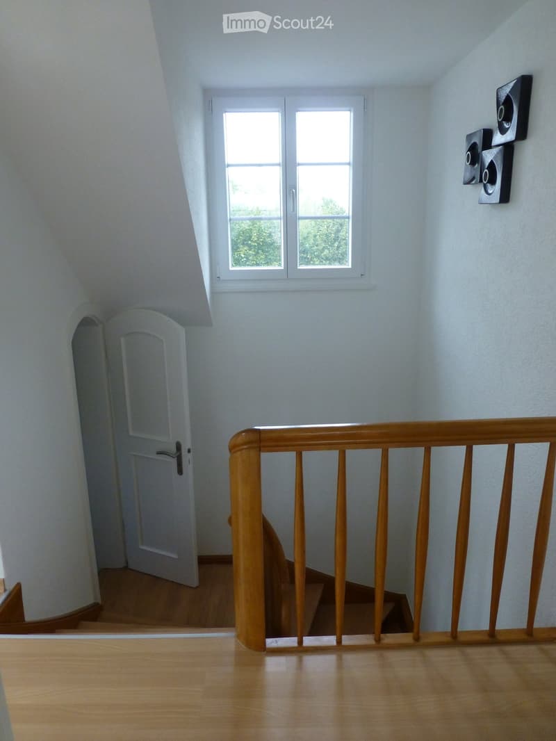 A narrow hallway with wooden flooring, white walls, and a wooden railing leading to a room. A white door is visible on the left, and a window with double panes is on the right wall.