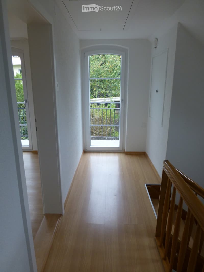 Bright hallway with wooden flooring, white walls, and a staircase leading upwards. A large window at the end provides natural light and a view of greenery outside.