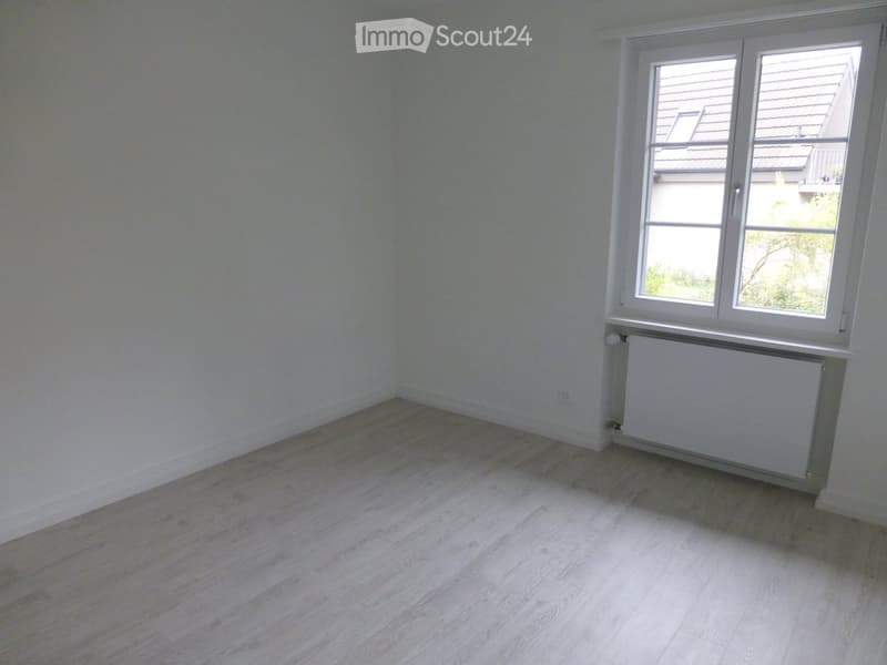 Empty room with light-colored wooden flooring and white walls, featuring a large window with white frames and a radiator below it.