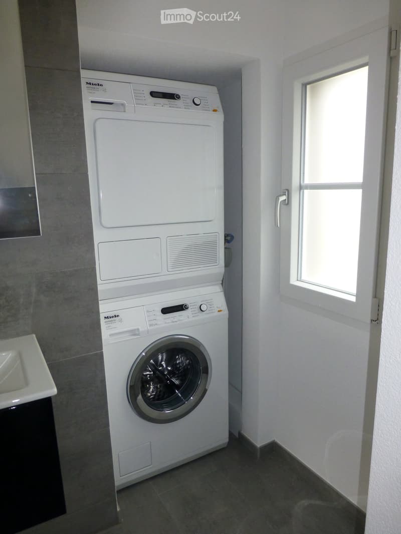 Bathroom with a white washing machine and dryer unit, a small sink, and a window letting in natural light.