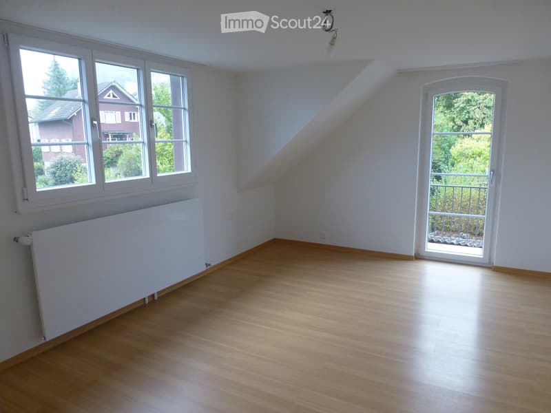 Empty room with wooden flooring, white walls, large windows, and a radiator. A sliding door leads to a balcony or terrace area.