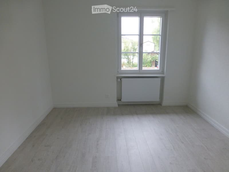 Empty room with light wooden flooring, white walls, and a single window with white frame. A radiator is visible below the window.