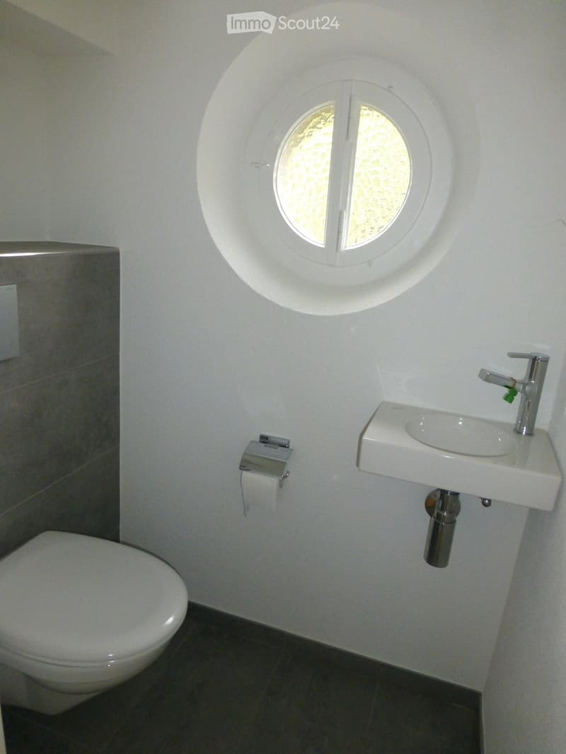 Modern bathroom with a toilet, wall-mounted sink, chrome faucet, and toilet paper holder. Features a round window with frosted glass above the sink.
