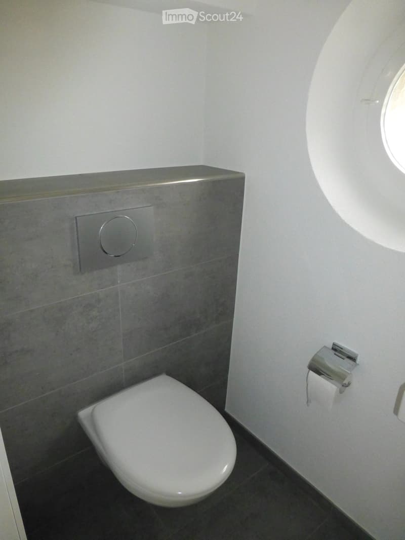 Modern bathroom with a white toilet, dark tiled floor, grey tiled wall, and a round window providing natural light.