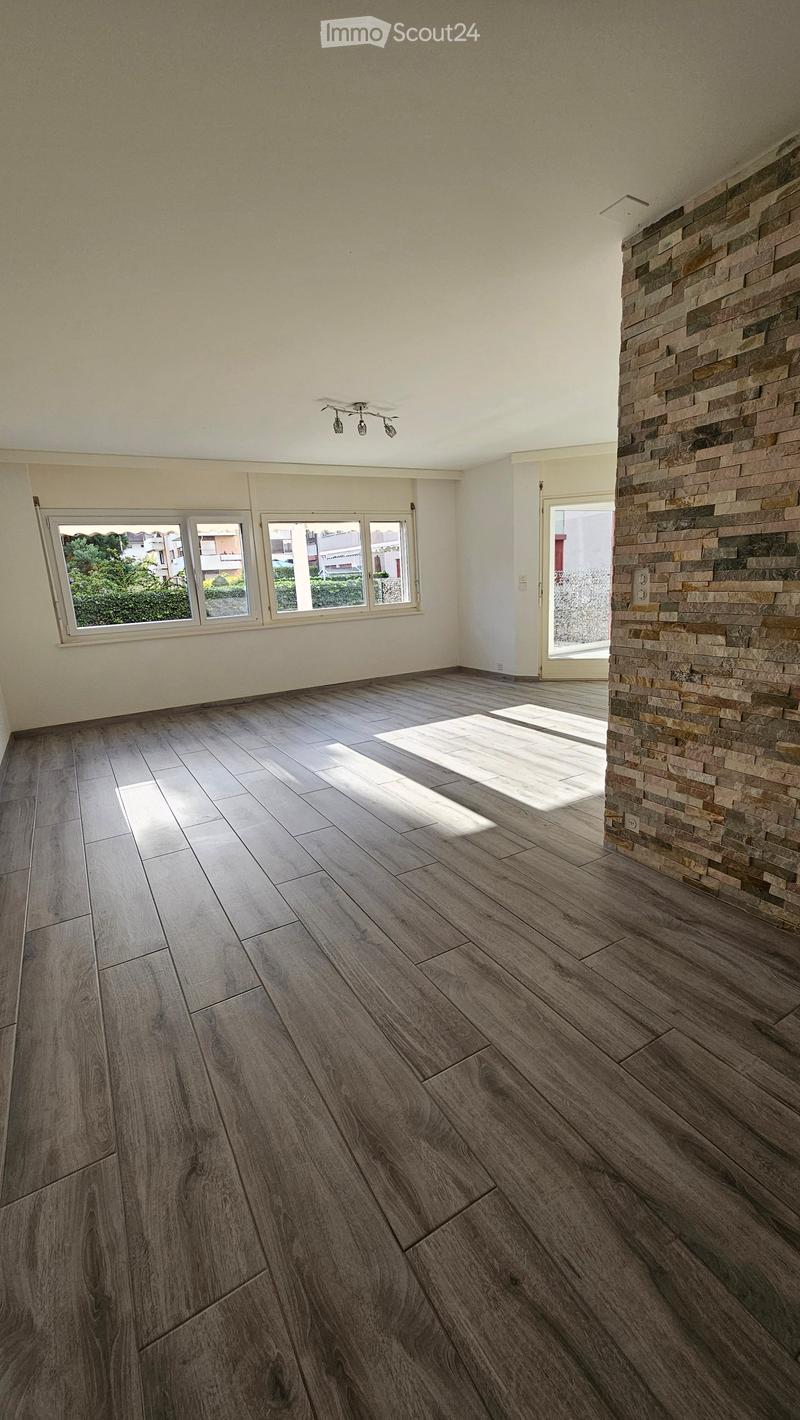 Empty room with wooden flooring, large windows, and a stone accent wall. No furniture or equipment present.