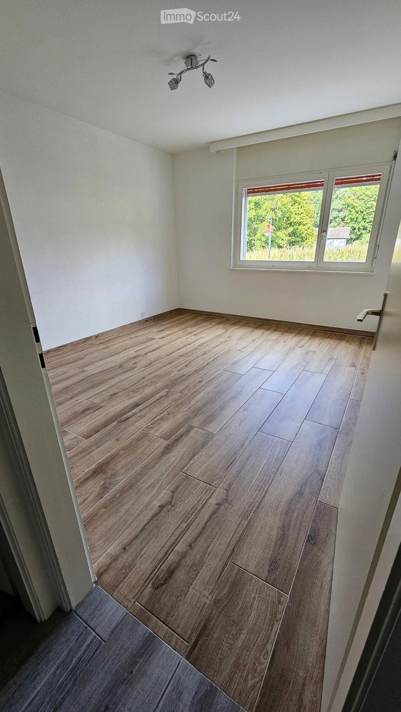 Empty room with wooden flooring, white walls, and a large window with a view of greenery outside.