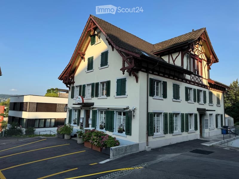 Two-story traditional building with white walls, green shutters, and a tiled roof. The ground floor features a restaurant sign and flower boxes, with a modern adjacent building and parking area.