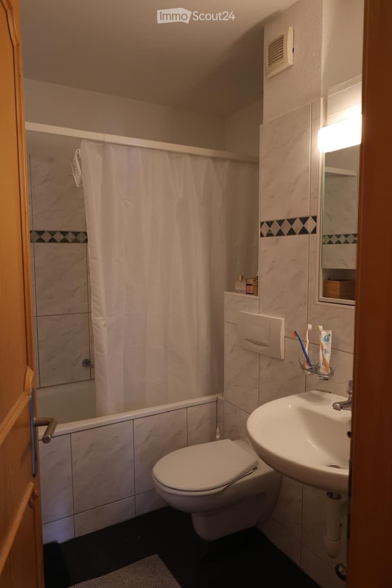 Modern bathroom with white tiles, a toilet, a sink with a mirror, a shower with a curtain, and a small shelf with toiletries.