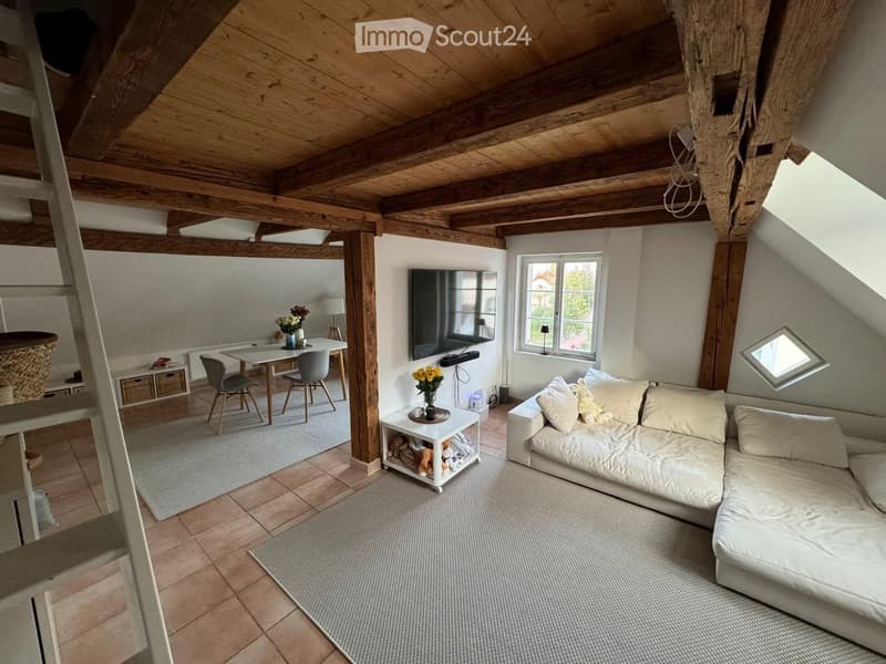 Cozy attic living space with exposed wooden beams, a white sectional sofa, a coffee table, a dining area with a table and chairs, and a television mounted on the wall.
