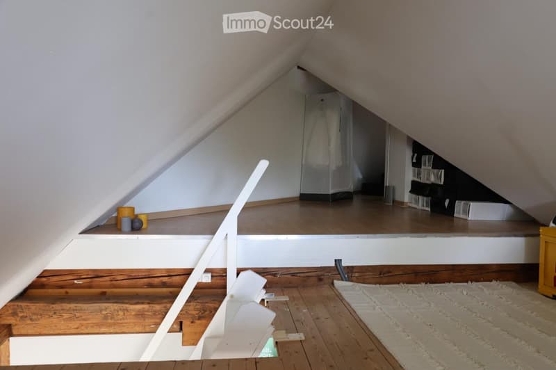 An attic space with wooden floors, slanted ceilings, and a small platform area. The lower level has a wooden staircase leading up, and the upper platform includes storage bins and a few decorative items like candles and a small globe.