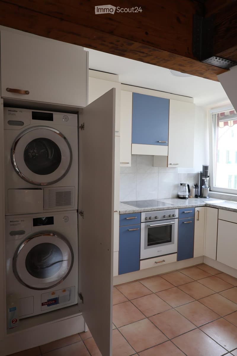 Modern kitchen with white and blue cabinets, tiled floor, and stainless steel appliances including an oven and washing machine. A coffee maker and cup are on the counter.
