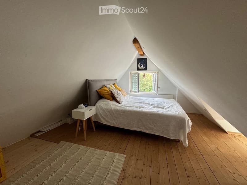 Cozy attic bedroom with wooden floors, slanted ceilings, a large window, a bed with pillows, a nightstand, and a rug.