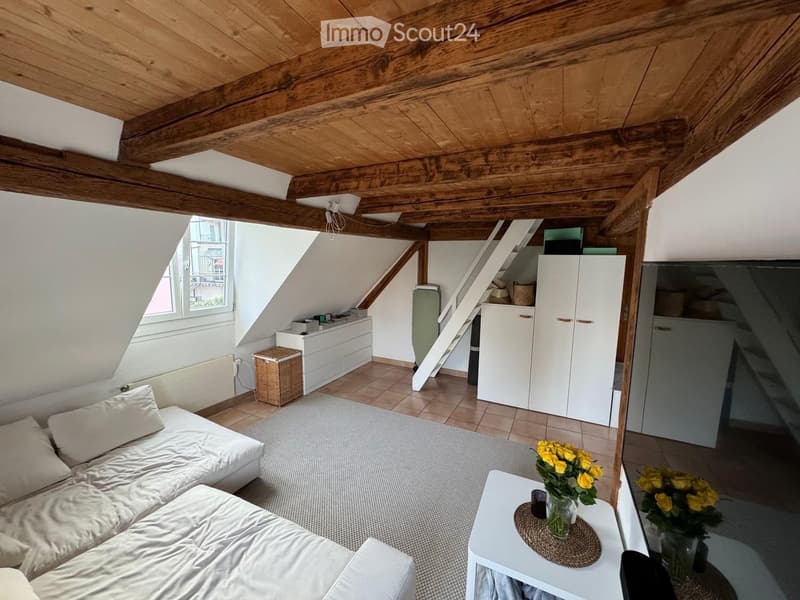 Loft-style living room with exposed wooden beams, white sofa, coffee table with flowers, white cabinet, wicker basket, and a staircase leading to another level.