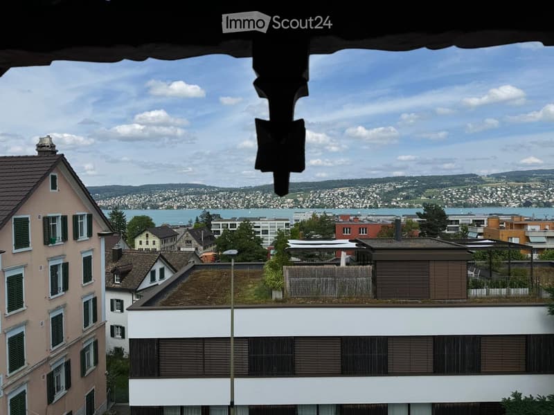 View from a balcony showing a cityscape with houses, a lake, and distant hills under a partly cloudy sky.
