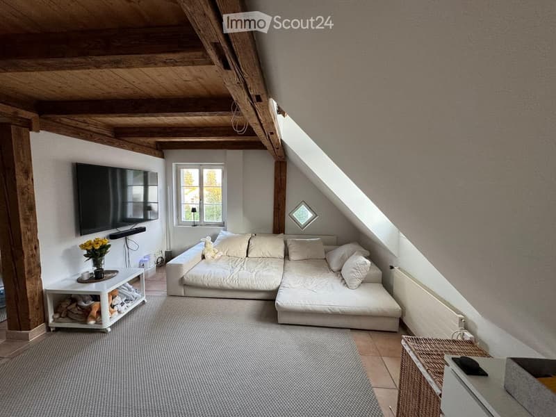 Cozy attic living room with exposed wooden beams, a large sectional sofa, a flat-screen TV, a small white side table with a vase of yellow flowers, and a wicker basket on the floor.