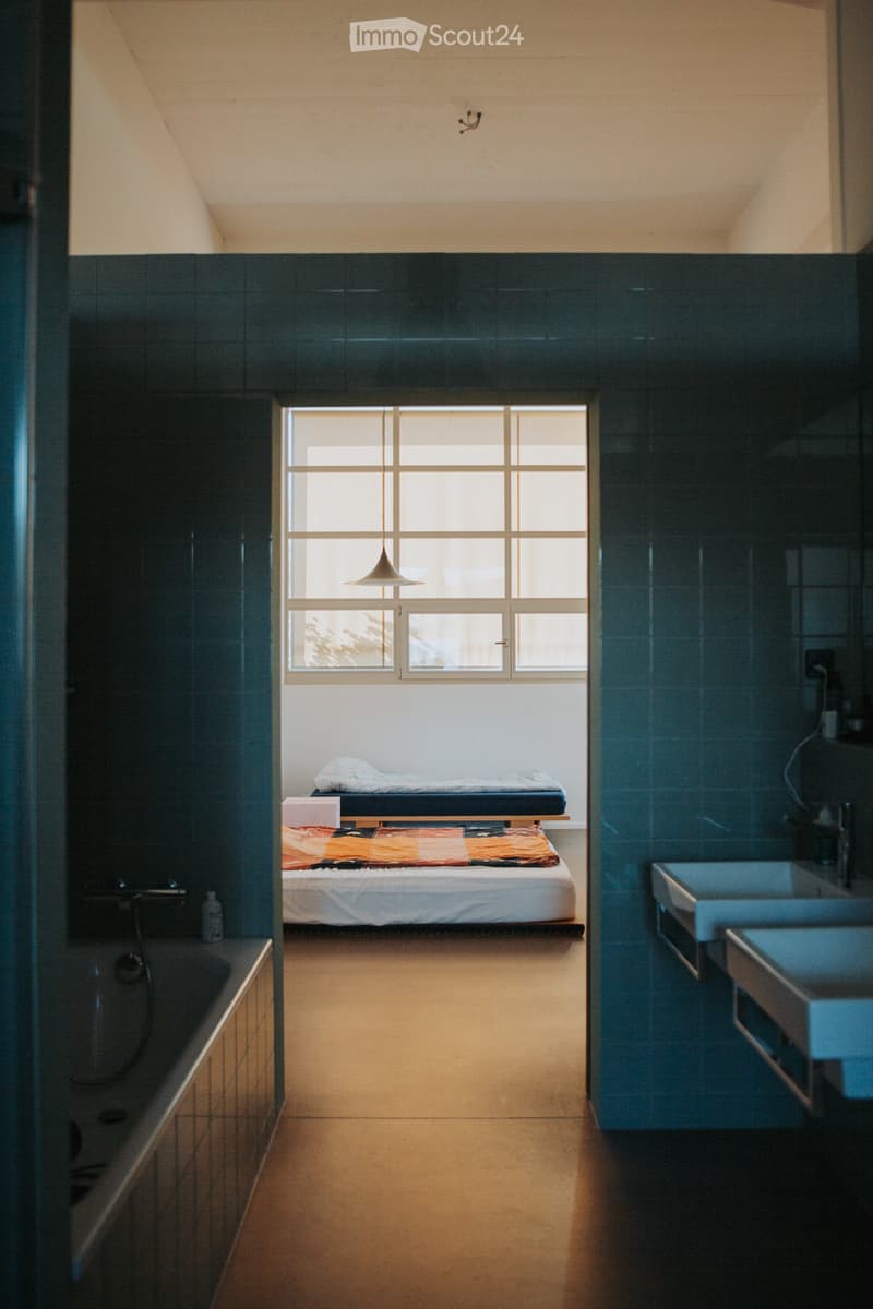 Modern bedroom with a bed, sink, and bathtub, featuring dark tiled walls and a large window letting in natural light.
