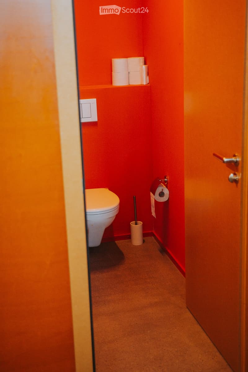 Small bathroom with red walls, white toilet, toilet paper holder, toilet brush, and a shelf with extra toilet paper rolls.