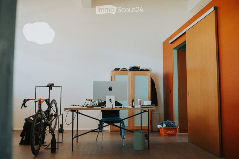 A furnished room with a wooden floor, a modern table with a laptop, a bicycle on a stand, a cabinet with glass doors, and a hanging cloud-shaped lamp.