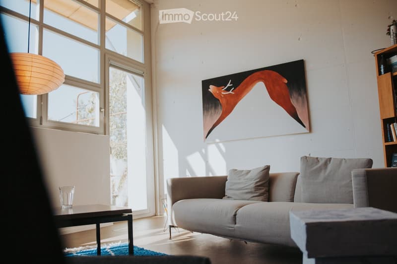 Modern living room with large windows, grey sofa, ottoman, coffee table, pendant light, bookshelf, and abstract painting.