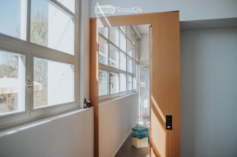 Bright hallway with large windows, light-colored walls, and a small stool in the corner.