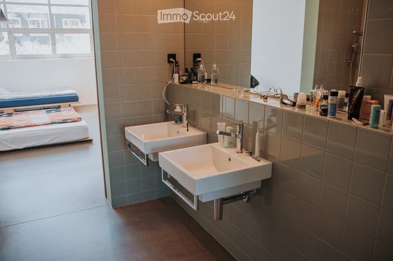 Modern bathroom with two white sinks, mirrors, and personal care items on the counter. The room features tiled walls and a large window letting in natural light.