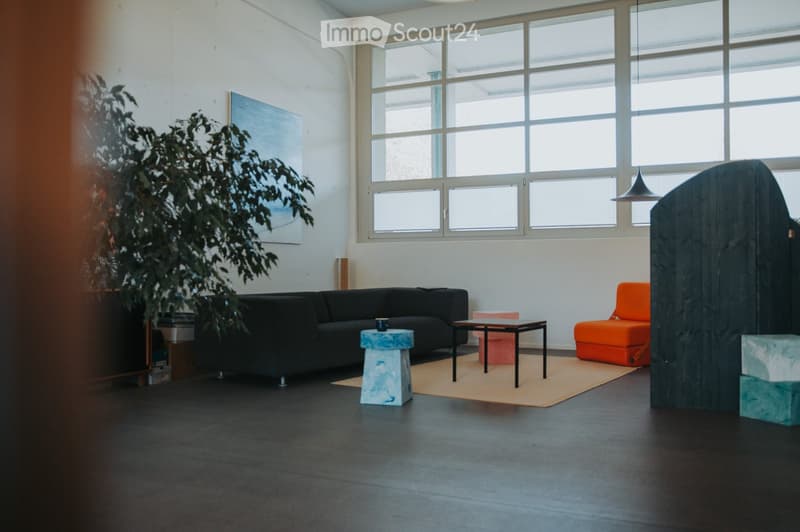 Modern living room with large windows, black sofa, orange chair, small tables, and a plant. The room features a dark floor and white walls.