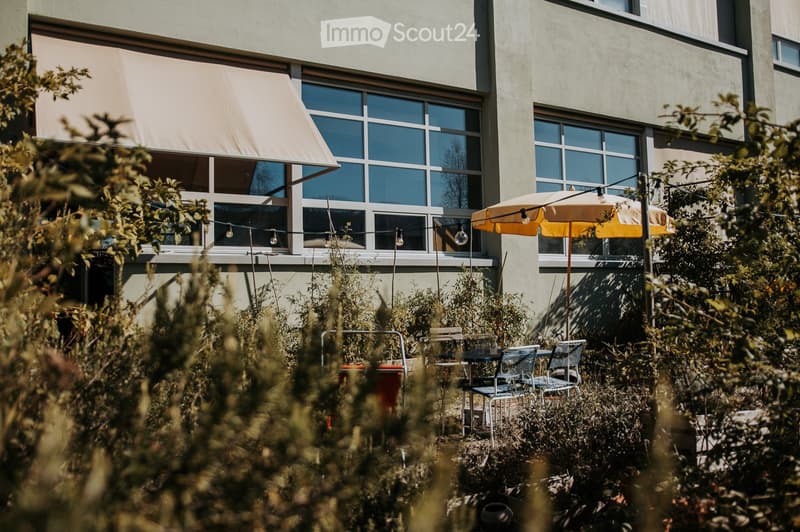 Outdoor seating area with a table, chairs, and an umbrella near a building with large windows and a covered awning.