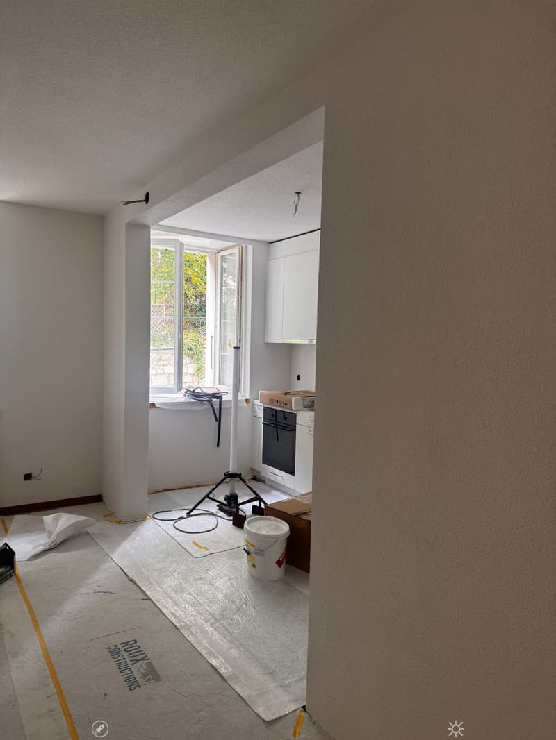 Empty room with unfinished walls and floor, featuring a small kitchen area with cabinets and an oven, a tripod, a bucket, and some construction materials.