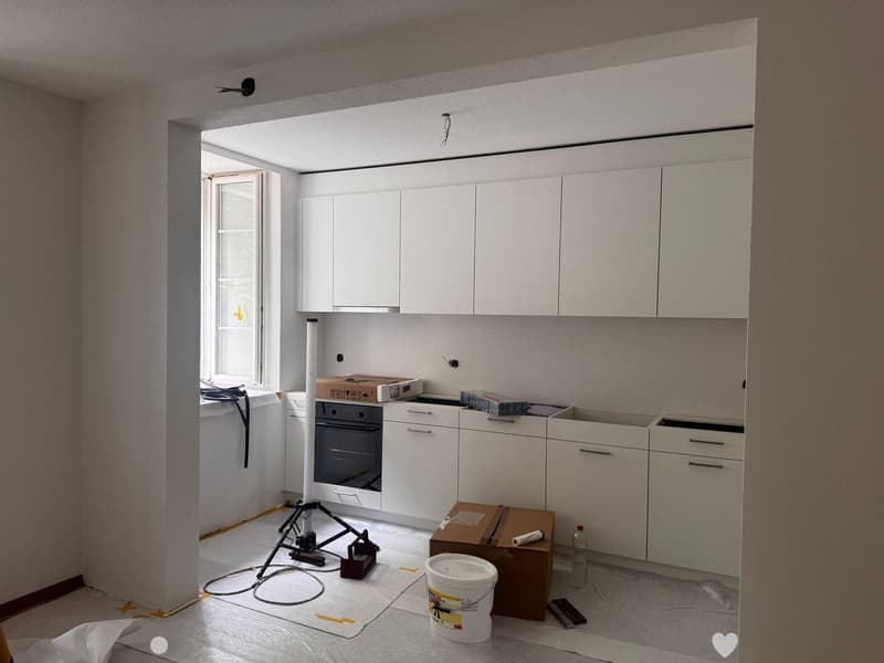 Empty kitchen with white cabinets, a window, and construction materials on the floor including a bucket, boxes, and a tripod.