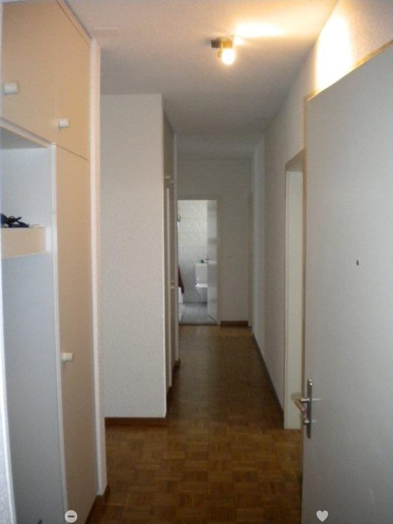 A narrow hallway with wooden flooring, white walls, and a single ceiling light. A door on the right leads to a room, and a cabinet is visible on the left side.