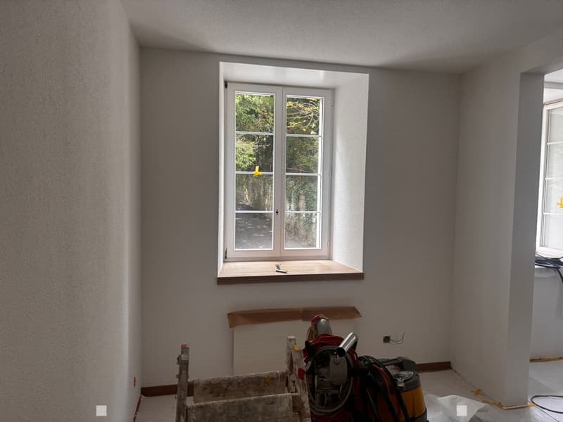 Empty room with unfinished walls, a window with a wooden sill, and construction tools and materials scattered on the floor.