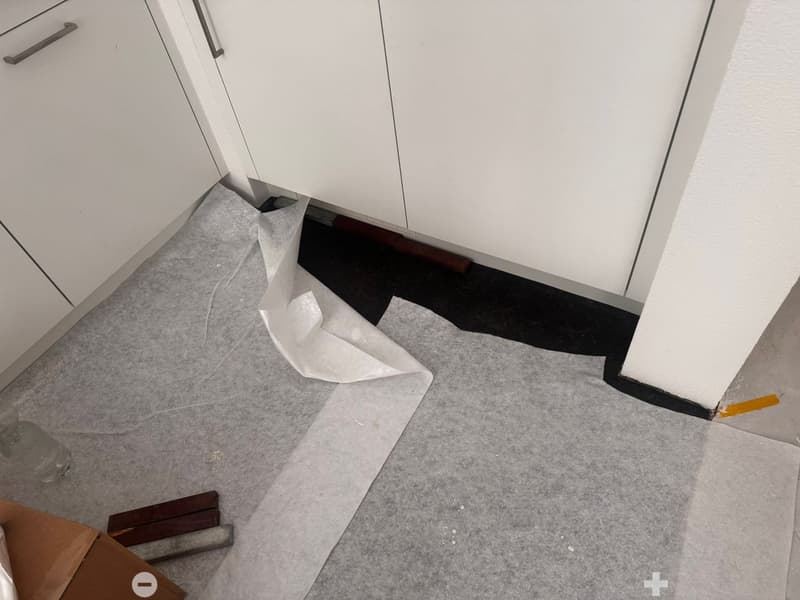 Empty room with grey carpet being installed, showing exposed subflooring and protective paper under the carpet. No furniture or appliances present.
