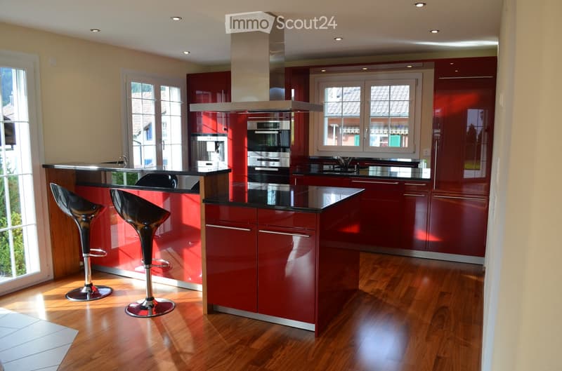 modern kitchen with red cabinets, black countertops, stainless steel appliances, wooden floor, two black bar stools, large windows