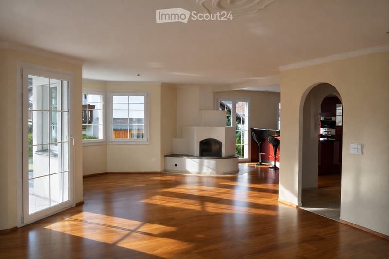 Bright living room with wooden flooring, large windows, fireplace, and two stools near a kitchen area.