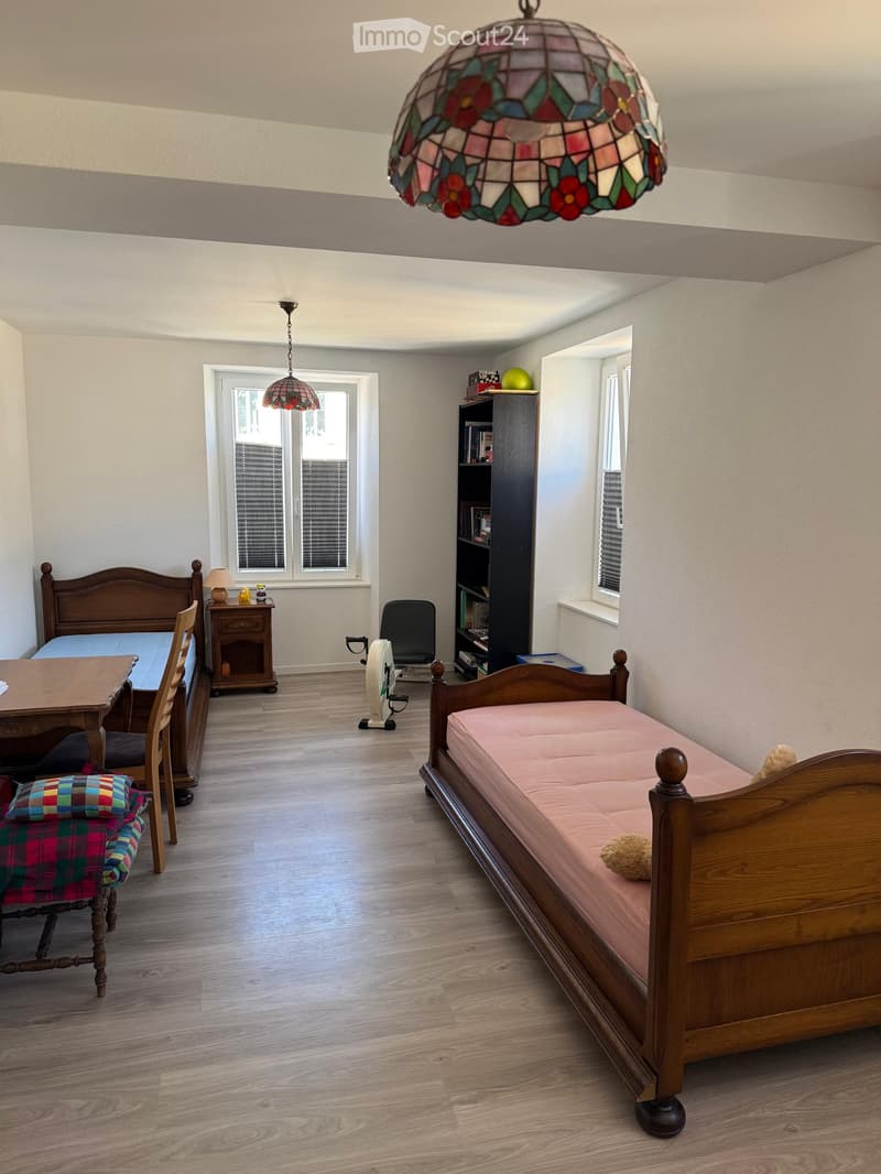 Bright bedroom with wooden flooring, a double bed with pink bedding, a wooden chair, a small table, a bookshelf, and a hanging stained-glass lamp.