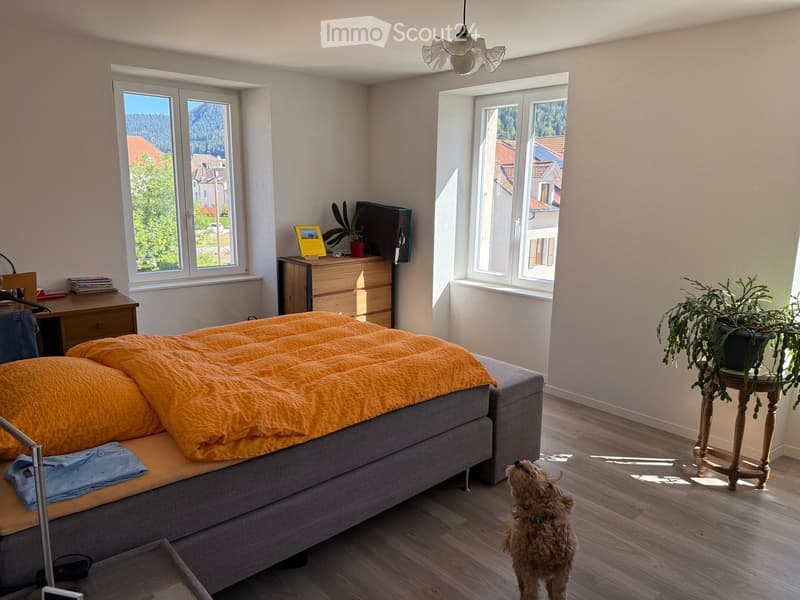 Bright bedroom with wooden flooring, large windows offering a scenic view, a grey bed with orange bedding, a nightstand with a plant, a small table with another plant, and a dog standing near the bed.