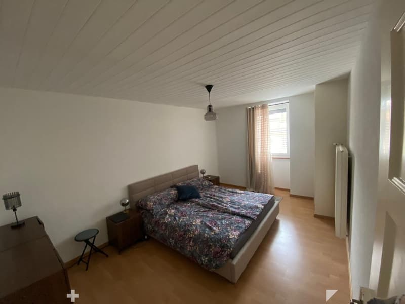 Cozy bedroom with wooden flooring, slanted ceiling, and large window with curtains. Features a bed with floral bedding, nightstands, and a hanging light fixture.
