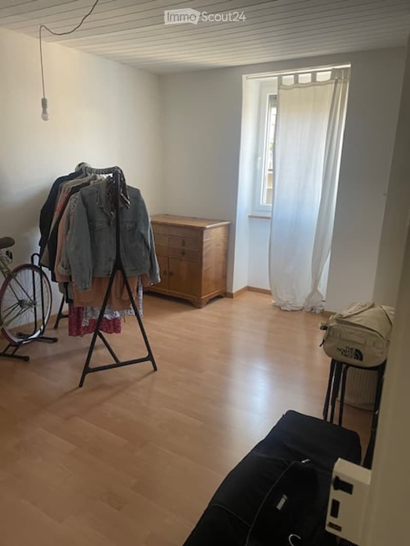 The room features wooden flooring, white walls, and a large window with white curtains. A clothing rack with several garments is positioned near the center, alongside a wooden wardrobe. A small stool and a backpack are placed near the window, and a suitca