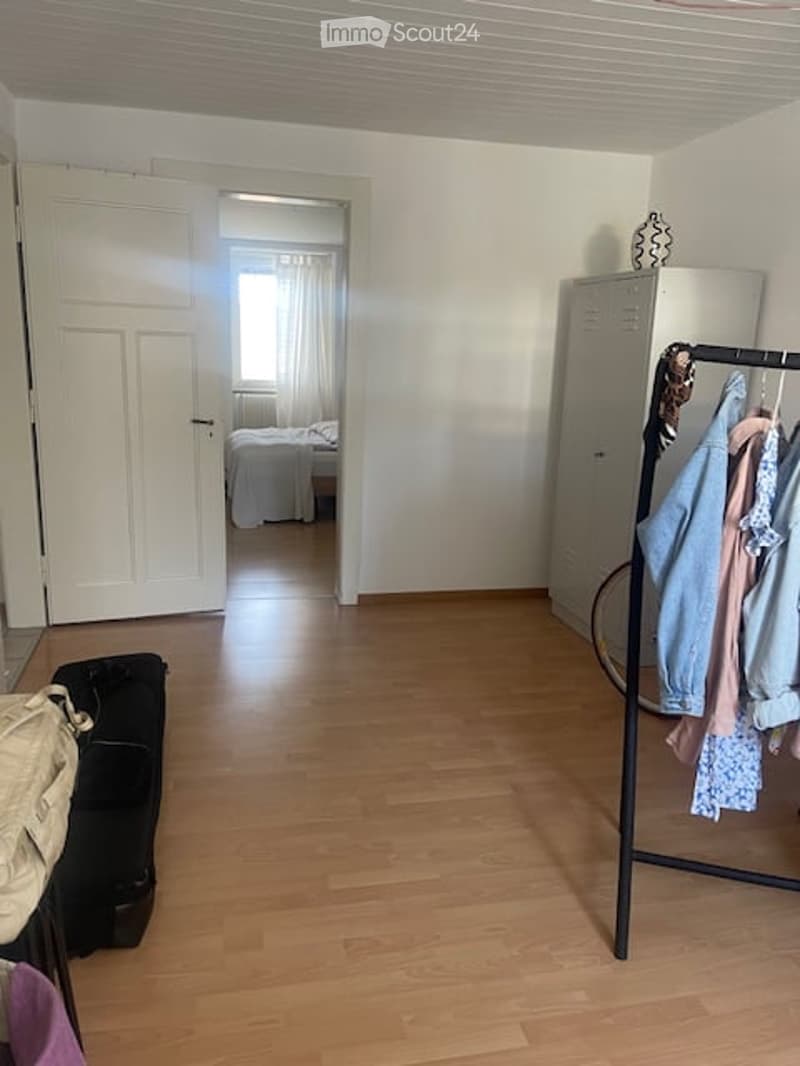 Empty room with wooden flooring, white walls, and a partially open door leading to another room. A clothing rack with garments and a suitcase are visible.