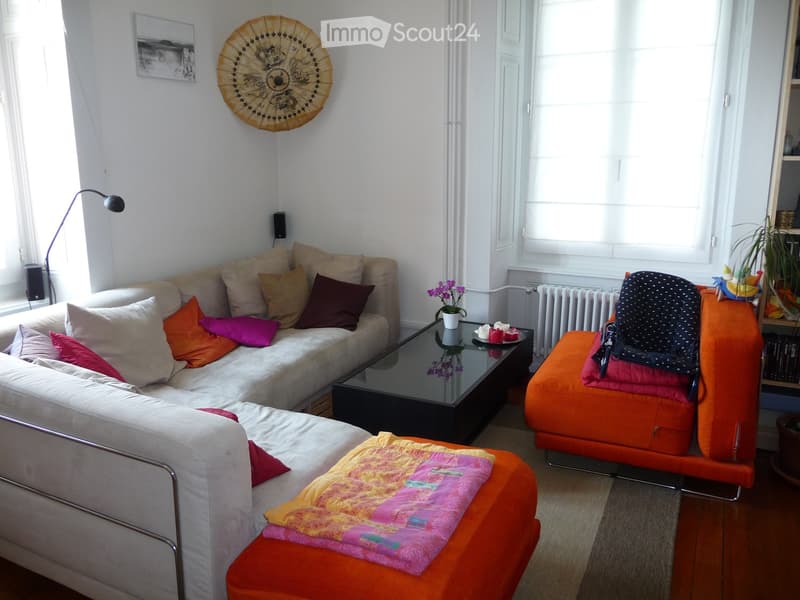 Modern living room with a white sectional sofa, colorful cushions, a glass coffee table with a flower vase, an orange ottoman, and a floor lamp. The room features large windows, a radiator, and a bookshelf with decorative items.