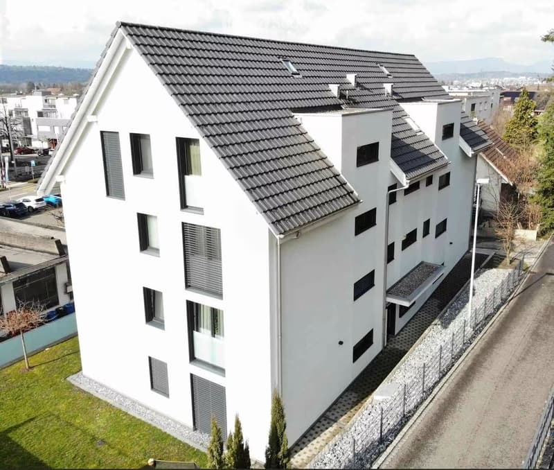 A modern white building with multiple floors and several windows. Located on an elevated floor, the house has a gray tiled roof and is surrounded by greenery and paved walkways.