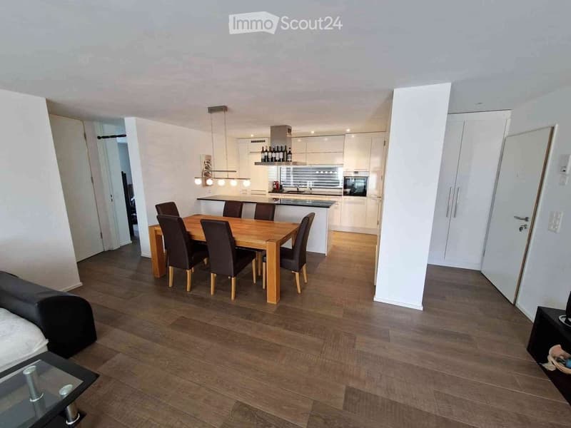 Dining room with wooden dining table, black chairs, modern kitchen with island, wooden floor, white walls, white cupboards, dining table, lights, microwave, and kitchen cabinets.