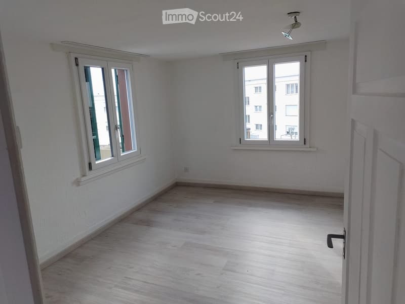 Empty room with light wooden flooring, white walls, and two large windows with white frames. The room has a simple, clean design with no furniture or decorations.