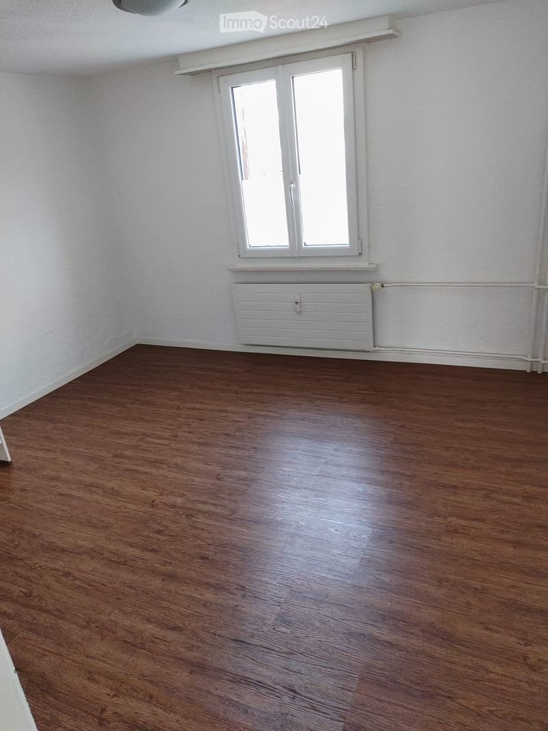 Empty room with wooden flooring, white walls, a window with white frame, and a white radiator below the window.