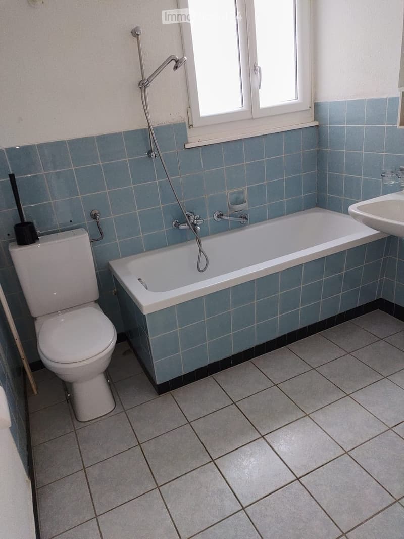 Bathroom with blue tiled walls, a bathtub, toilet, sink, and a window above the bathtub.