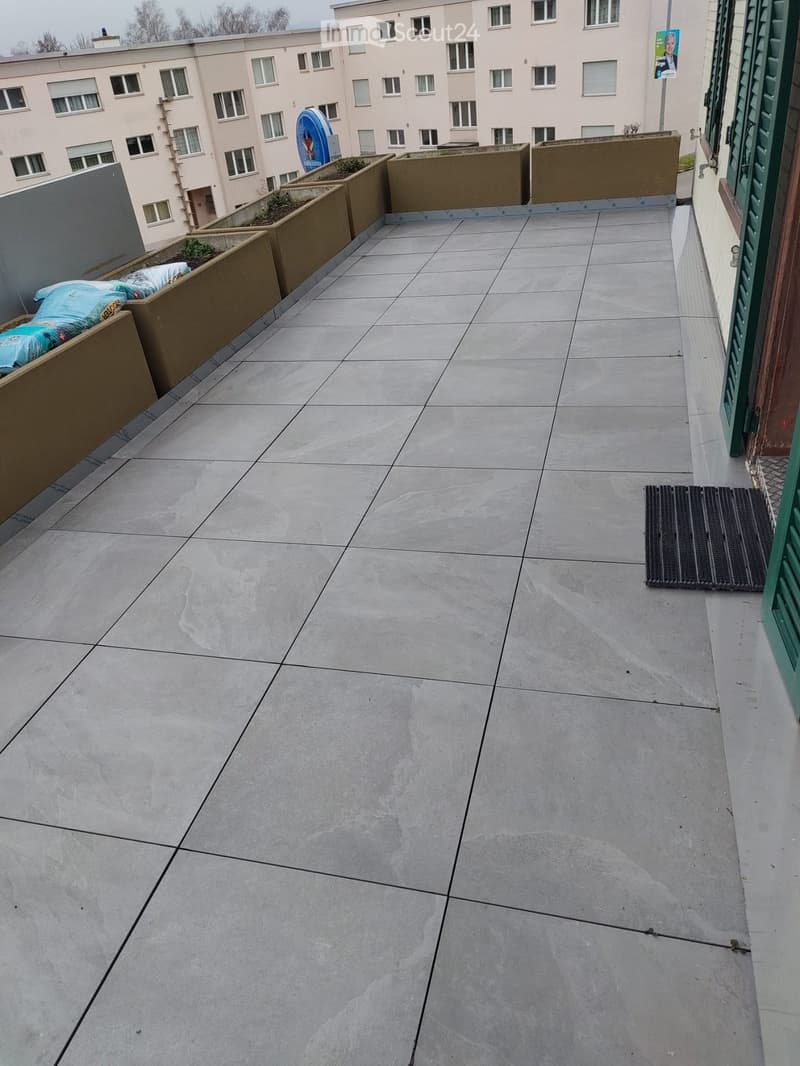 Empty balcony with tiled floor, raised planters, and a view of a multi-story building in the background.
