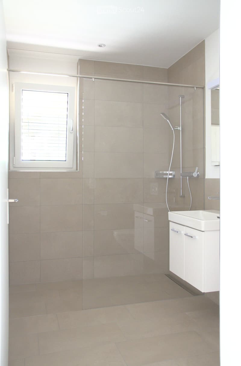 Modern bathroom with beige tiles, a white sink with cabinet, a glass shower enclosure, and a window with blinds.