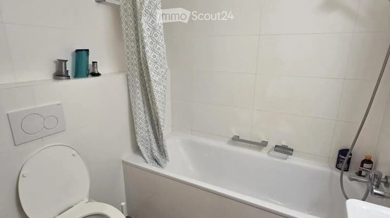 Bathroom with a toilet, bathtub, and a shower curtain. The bathtub has a handheld shower attachment, and there are various toiletries on the shelf above the bathtub.