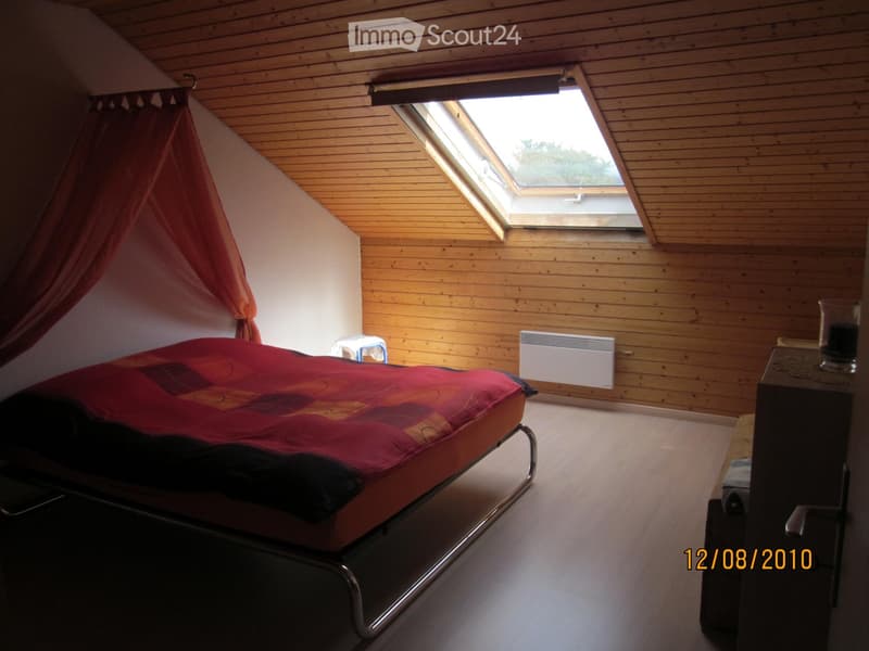 Cozy attic bedroom with wooden walls and ceiling, skylight window, bed with red and orange bedding, nightstand, and small stool.